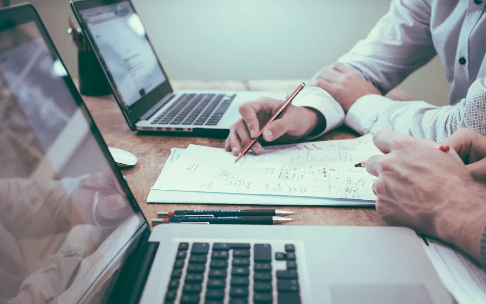 Two people at a desk marking up a printed document with pencils, laptops open beside them